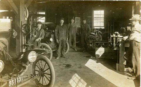 1915 Ford Model T - A group of men work in a garage, with a 1915 Ford Model T visible in the foreground and other vehicles in the background.