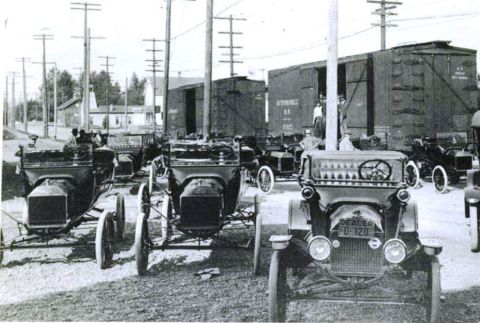 1915 Ford Model T - A group of vintage automobiles are lined up outdoors, with boxcars visible in the background.