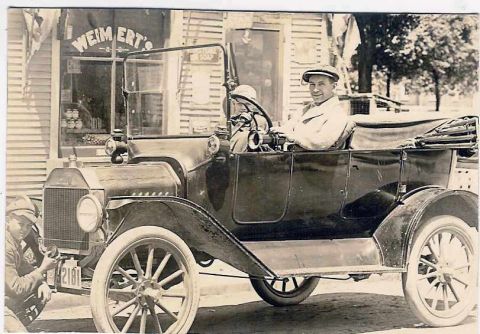 1915 Ford Model T - A man in a cap drives a 1915 Ford Model T touring car parked in front of a store with "WEIMERT'S" visible on the window.