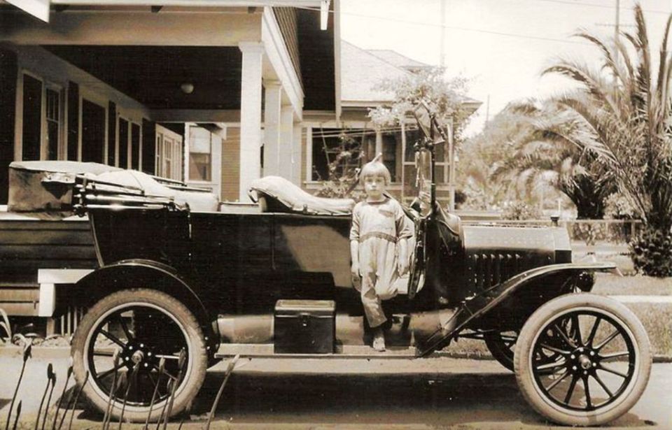 Boy in a light-colored outfit stands on the running board of a dark-colored touring car. A house with a porch is behind them