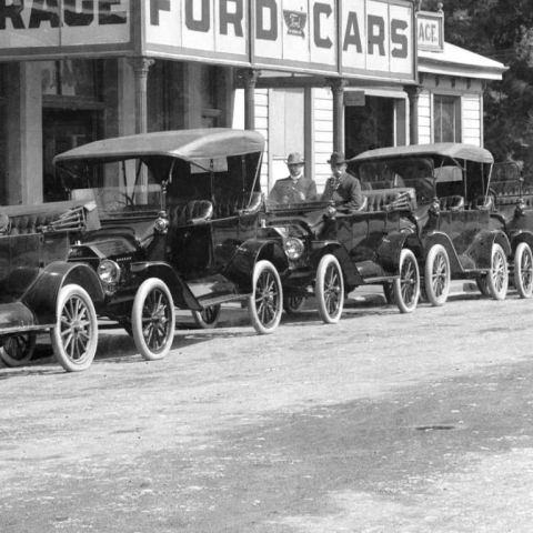 1915 Ford Model T - A row of Ford Model T touring cars are lined up in front of a dealership with 'Ford Cars' visible on the signage.