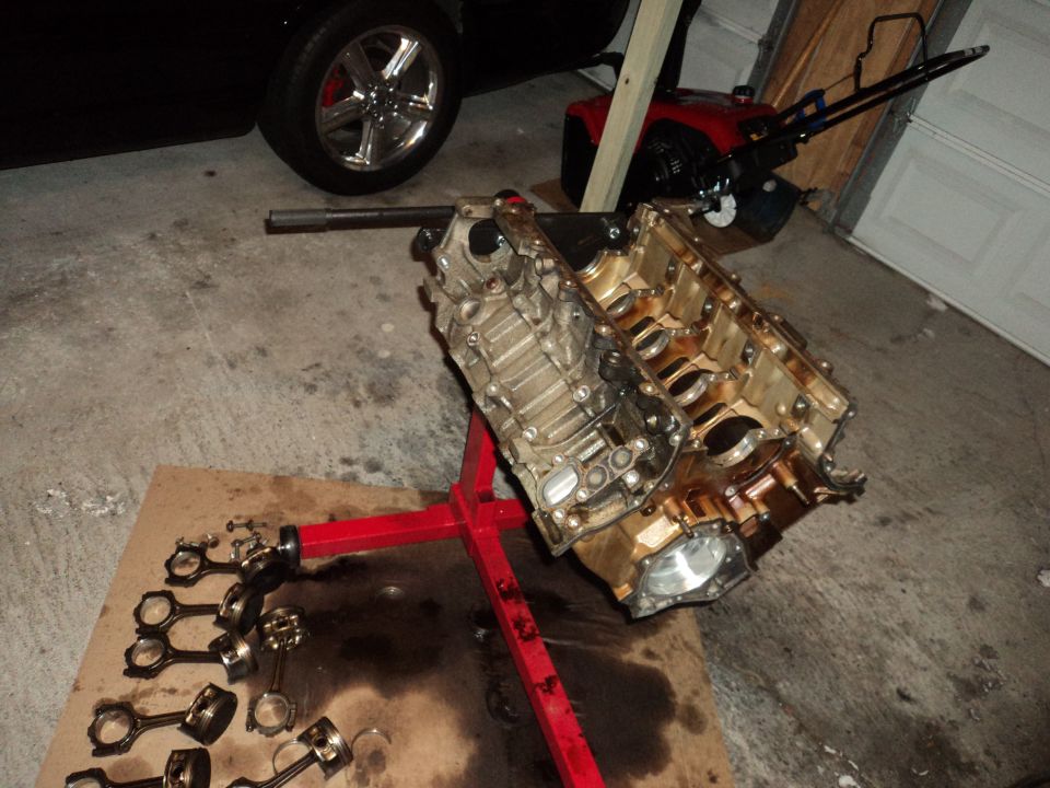 Engine on a red stand, with pistons and connecting rods laid out on cardboard. A black car wheel and a snowblower are in the background