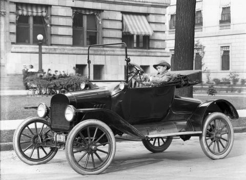 1915 Ford Model T - A young boy at the wheel of a 1915 Ford Model T touring car, parked on a street with a building in the background.