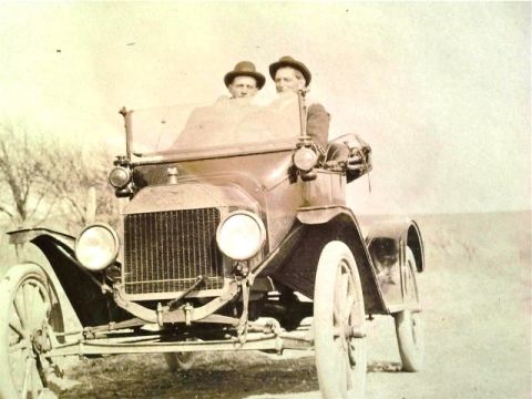 1915 Ford Model T - An early automobile with two occupants drives down a dirt road on a clear day.