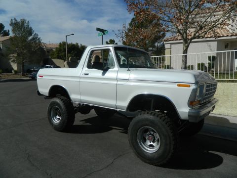 1979 Ford Bronco - This lifted white Ford Bronco is parked on a street with houses and trees in the background.