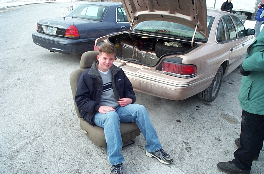 Man sits on a car seat with the trunk open of a 1991 Ford Crown Victoria LTD. Another Ford Crown Victoria is parked behind it