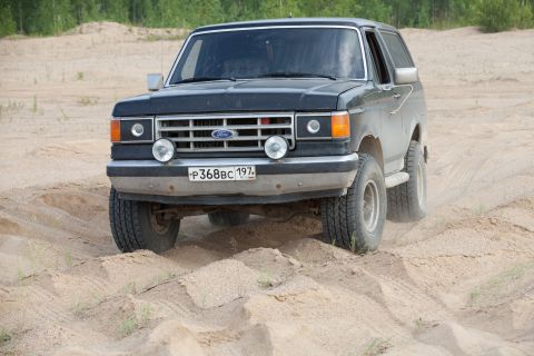 1987 Ford Bronco - A black 1987 Ford Bronco navigates a sandy terrain, kicking up dust.