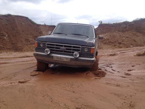 1987 Ford Bronco - A dark blue 1987 Ford Bronco navigates a muddy terrain, its front tires deeply embedded in the mire.