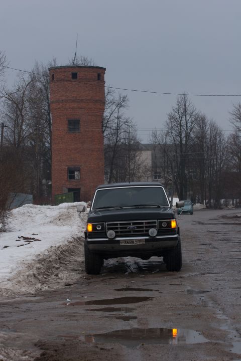 1987 Ford Bronco - A black 1987 Ford Bronco is seen from the front, driving on a wet, muddy road next to a snow-covered embankment.