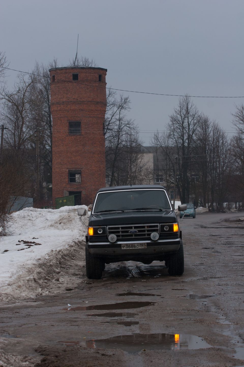 Black 1987 Ford Bronco front view, driving on a wet road with snow on the side