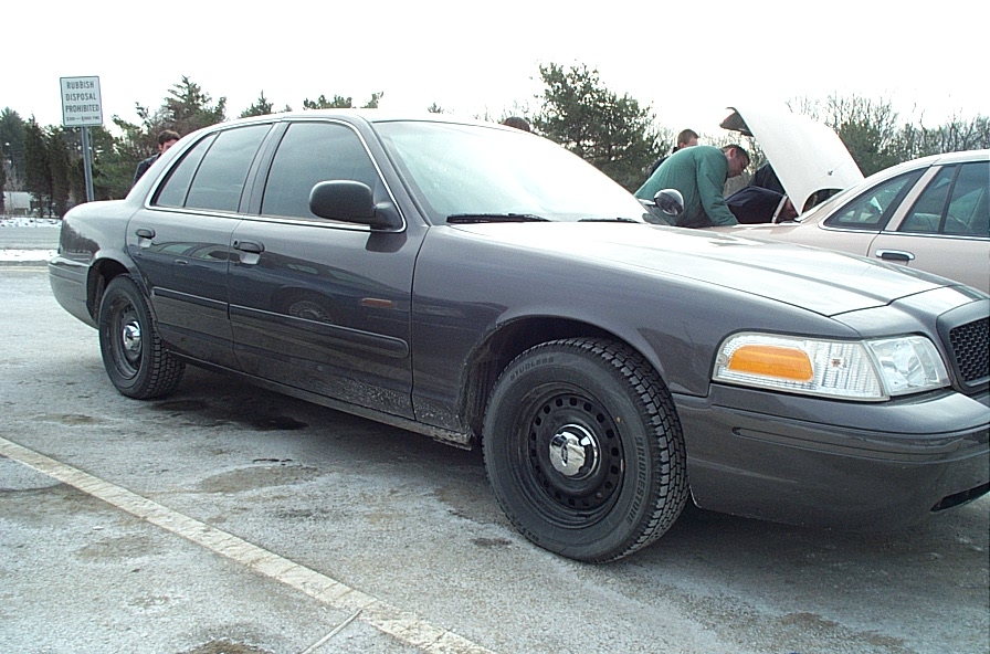 Dark gray sedan parked on asphalt, front passenger side visible. Hood is open, with people around