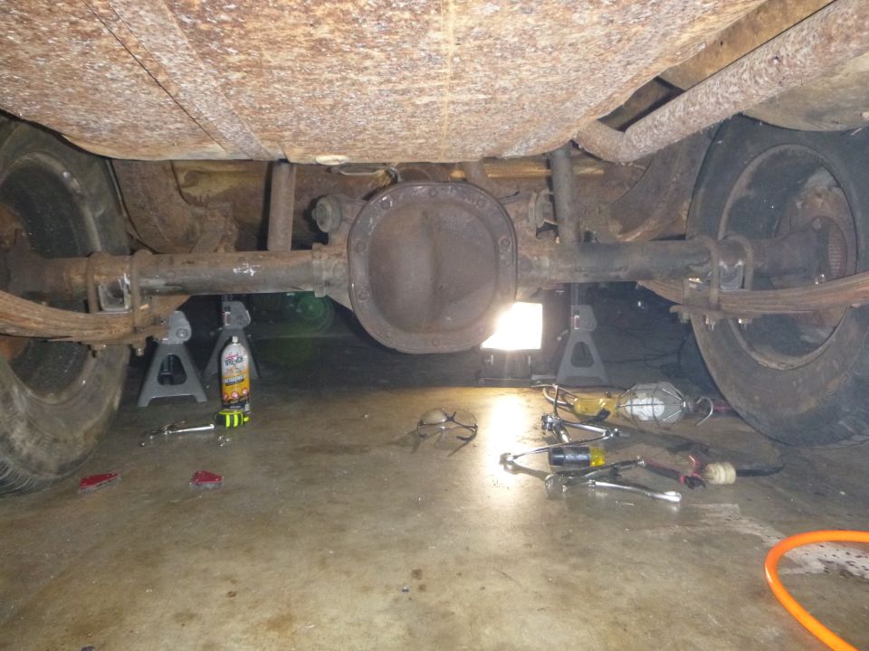 Underside view of a car's rear axle assembly supported by jack stands, with tools on the floor