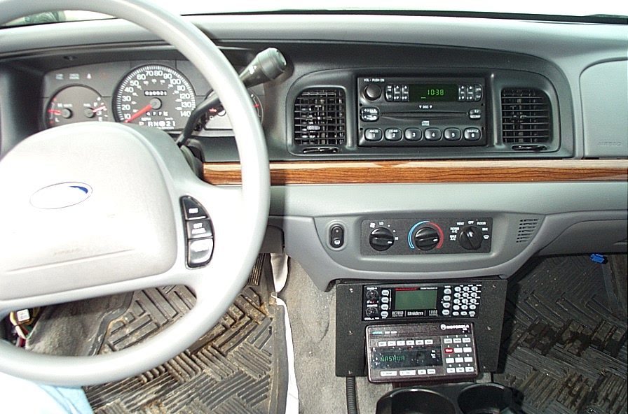 Interior view of a Ford Crown Victoria showing the steering wheel, dashboard, radio, and two police scanners mounted below the climate controls