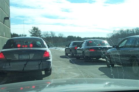1991 Ford LTD Crown Victoria - Multiple dark sedans queue up on a sunny day, with snow visible on the roadside.