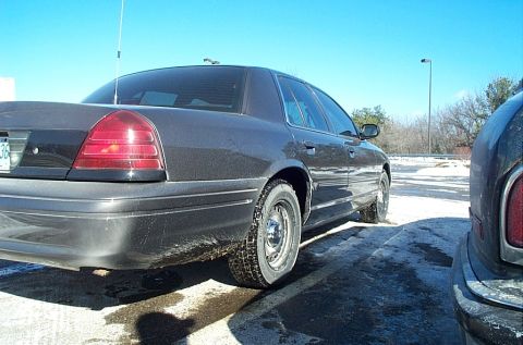 1991 Ford LTD Crown Victoria - A dark sedan is seen from the rear quarter panel in a snowy parking lot, with another vehicle partially visible on the right.