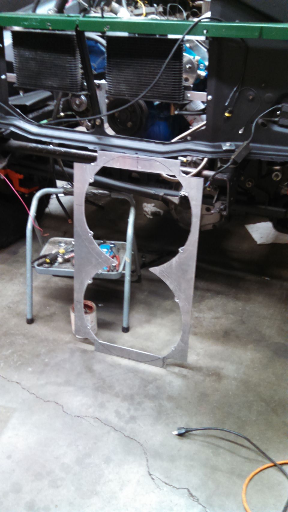 Custom aluminum fan shroud with two large circular cutouts, resting on the floor in front of a vehicle's engine bay