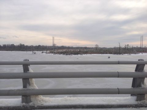1979 Ford Bronco - A frozen body of water stretches to the horizon under a cloudy sky, viewed from behind a metal bridge railing.