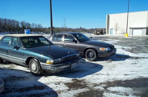 1991 Ford LTD Crown Victoria - A dark grey sedan is parked in a lot with patches of snow and ice, seen from a three-quarter front angle.