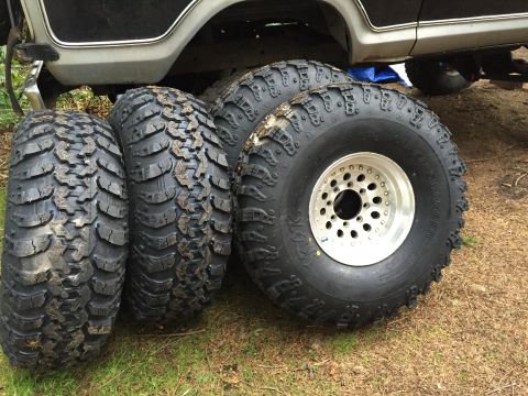 1971 Ford F-250 - Four aggressive off-road tires are displayed next to a single polished aluminum wheel, with the lower body of a vehicle visible in the back…