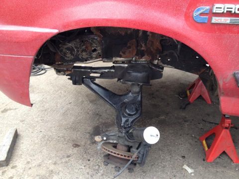 1989 Ford Bronco - Vehicle suspension parts are visible, with the body of a red vehicle above and jack stands supporting the frame.