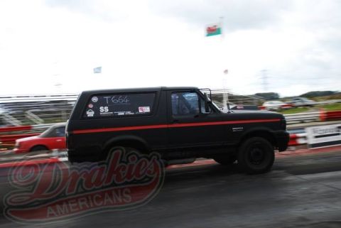 1989 Ford Bronco - A black Ford Bronco with a red stripe races down a track, with another car blurred in the background.