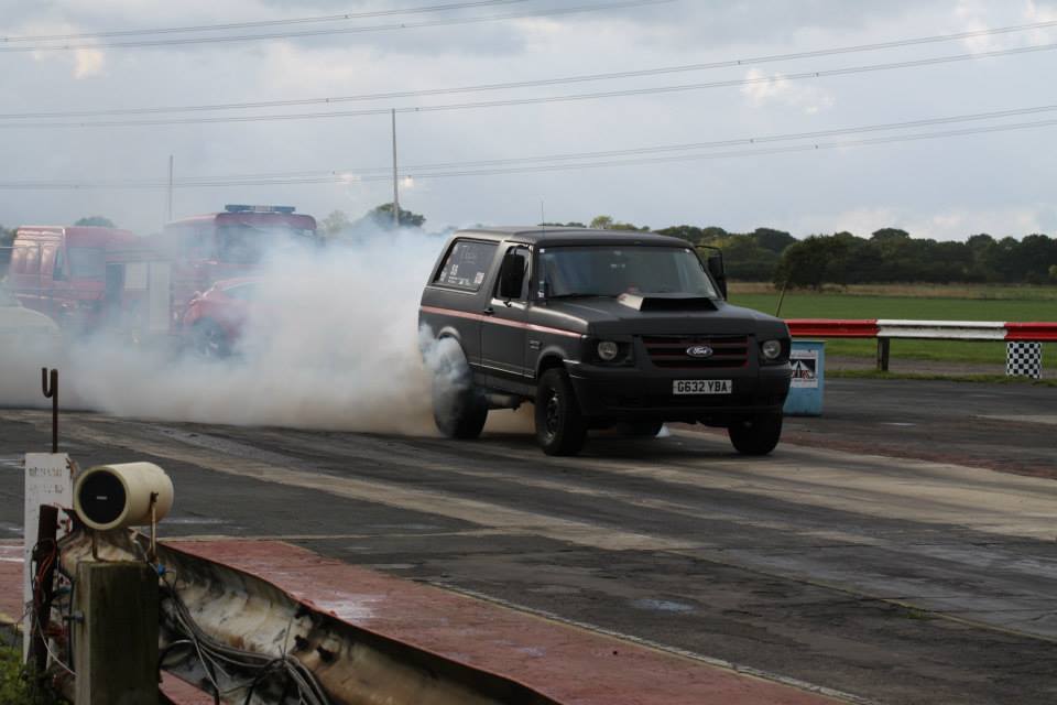 1989 Ford Bronco doing a burnout on a drag strip, with another vehicle in the background
