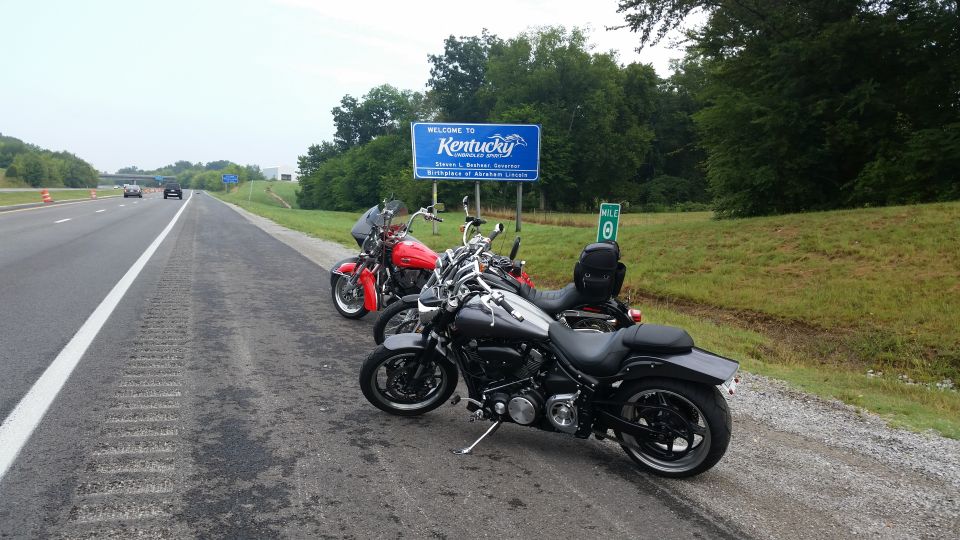Two motorcycles parked on highway shoulder near Kentucky welcome sign. One is black, the other red