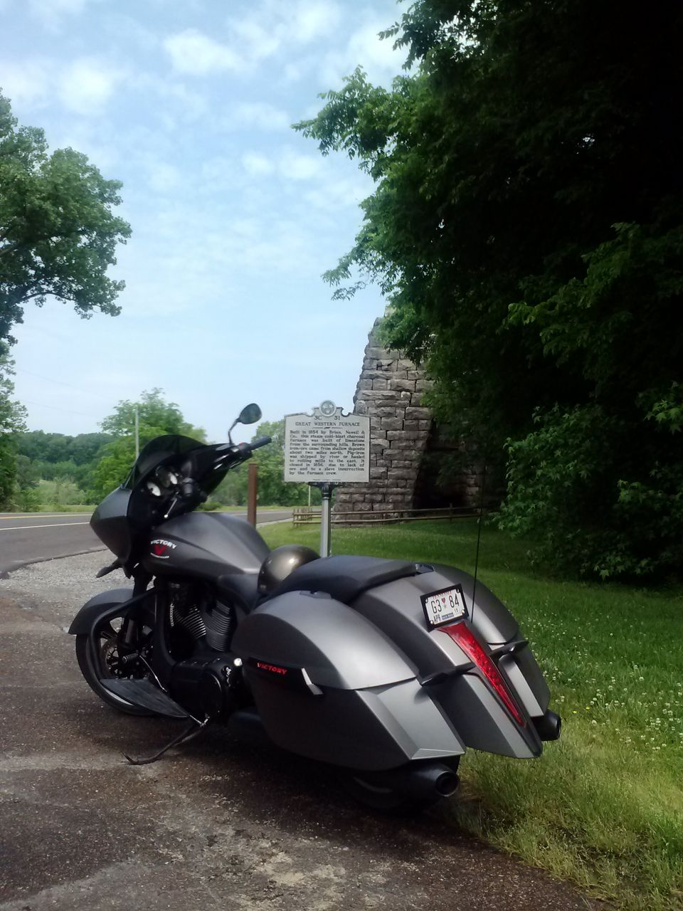 Gray Victory motorcycle parked on a paved area next to a grassy embankment with a stone structure and historical marker in the background