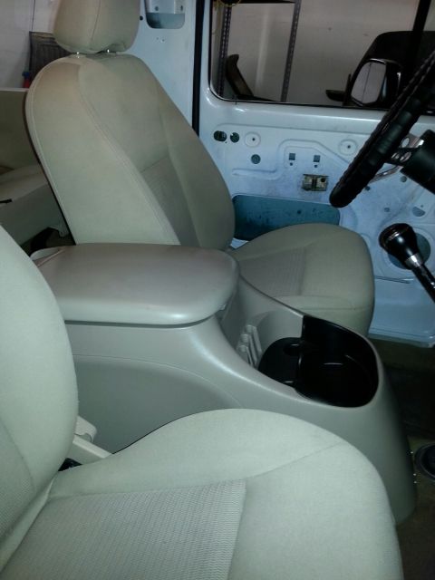 1979 Ford Bronco - The interior of a vehicle is shown, tan seats and a center console with a black cup holder insert.