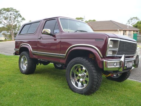 1985 Ford Bronco - This maroon 1985 Ford Bronco features chrome accents and custom wheels, parked on a grassy lawn.