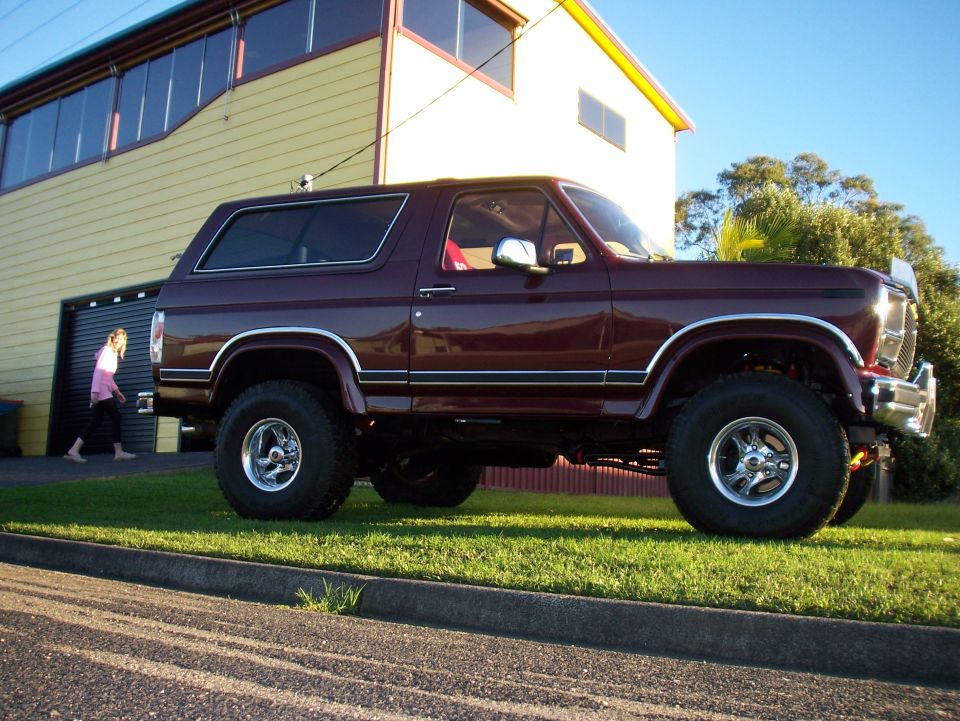 Maroon 1985 Ford Bronco parked on grass in front of a yellow building