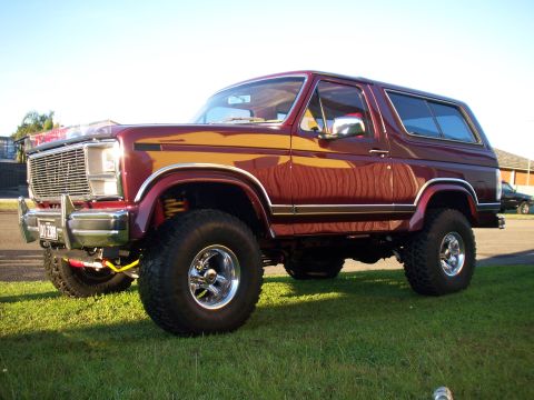 1985 Ford Bronco - This maroon Ford Bronco features a lifted suspension and chrome wheels, parked on a grassy area under a clear sky.