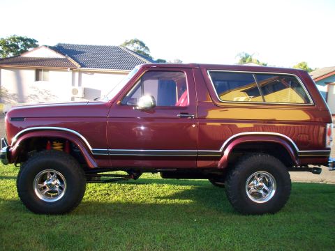 1985 Ford Bronco - This maroon 1985 Ford Bronco is shown from the side, parked on a grassy lawn in front of a house.