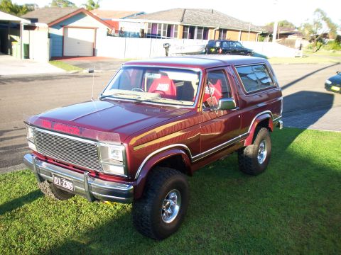 1985 Ford Bronco - Maroon Ford Bronco with red interior, lifted, chrome wheels, parked on grass.