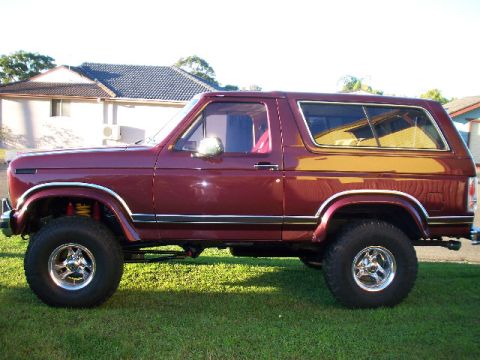 1985 Ford Bronco - This maroon Ford Bronco is shown from the side, parked on a grassy lawn with a house in the background.