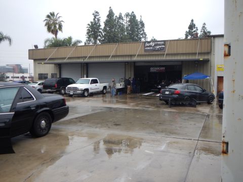 2003 Ford Crown Victoria - On a rainy day, several cars including a black sedan and white pickup sit on wet pavement outside an open dyno shop garage.