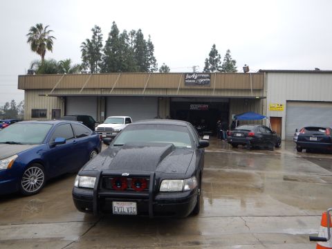 2003 Ford Crown Victoria - black sedan with a front brush guard and red-tinted accents, covered in rain droplets, parked outside an open garage on a wet day.