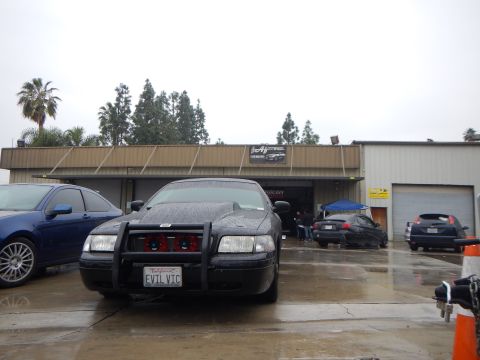 2003 Ford Crown Victoria - rain-speckled black sedan with a brush guard and EVIL VIC license plate faces an open dyno-shop bay on an overcast day.