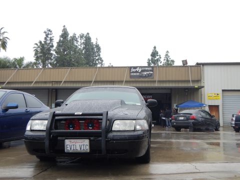 2003 Ford Crown Victoria - wet black sedan fitted with a front brush guard and an EVIL VIC plate sits before an open shop bay, a blue car alongside.
