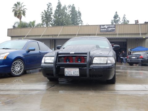 2003 Ford Crown Victoria - rain-covered black sedan with a brush guard and EVIL VIC license plate is parked next to a blue sedan outside a garage.