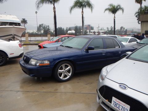 2003 Ford Crown Victoria - dark blue Mercury Marauder sedan with chrome wheels sits among other cars on a rain-soaked lot, a silver Ford and a boat nearby.