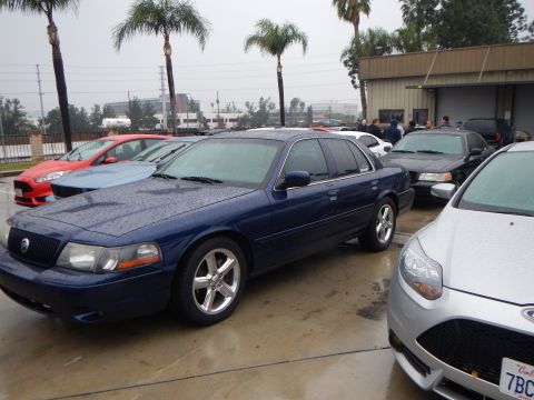 2003 Ford Crown Victoria - dark blue Mercury Marauder sedan parked next to a silver Ford Focus on a wet lot, with a red car and palm trees in the background.