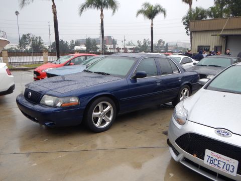 2003 Ford Crown Victoria - rain-beaded dark blue Mercury Marauder, its Mercury hood badge visible, parked beside a silver Ford on a wet lot with palm trees.