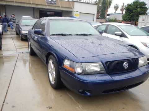 2003 Ford Crown Victoria - dark blue Mercury sedan with a chrome waterfall grille and Mercury emblem, wet with rain, parked in a row of cars at a shop.