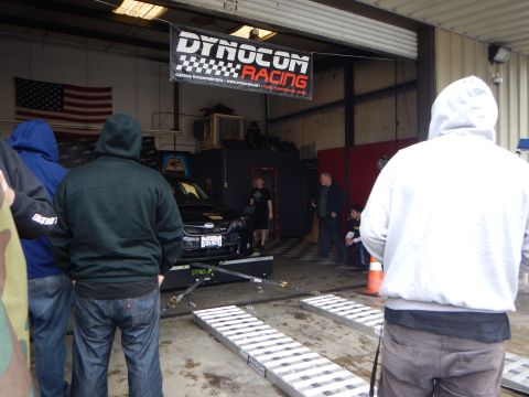 2003 Ford Crown Victoria - Spectators in hoodies stand watching a dark car strapped to a dyno inside a garage beneath a Dynocom Racing banner and US flag.