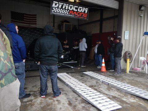 2003 Ford Crown Victoria - group of people watch a vehicle being dyno-tested in a garage, with aluminum ramps in the foreground and a Dynocom Racing banner above.