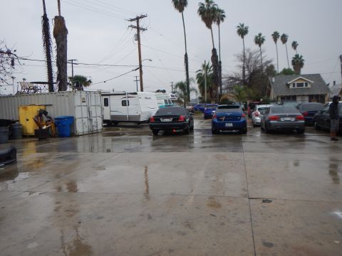 2003 Ford Crown Victoria - broad wet parking lot under gray skies holds several parked sedans, a white travel trailer and shipping containers among palm trees.