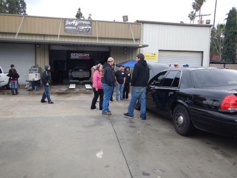 2003 Ford Crown Victoria - small crowd stands talking beside a black Ford Crown Victoria outside an open shop bay where a car sits on a dyno.