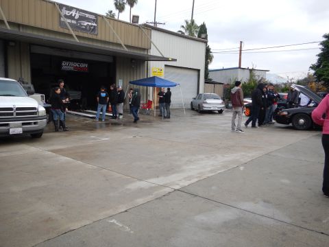 2003 Ford Crown Victoria - People mill about a wet shop lot with a black Crown Victoria, white pickup and blue canopy outside an open dyno-shop bay.