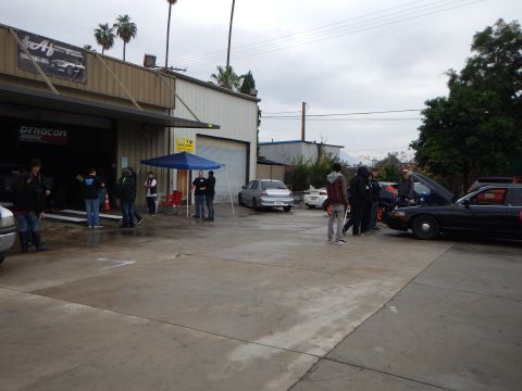 2003 Ford Crown Victoria - small crowd of people stands in the wet parking lot of an auto performance shop, with cars and a blue pop-up tent nearby under a gray sky.